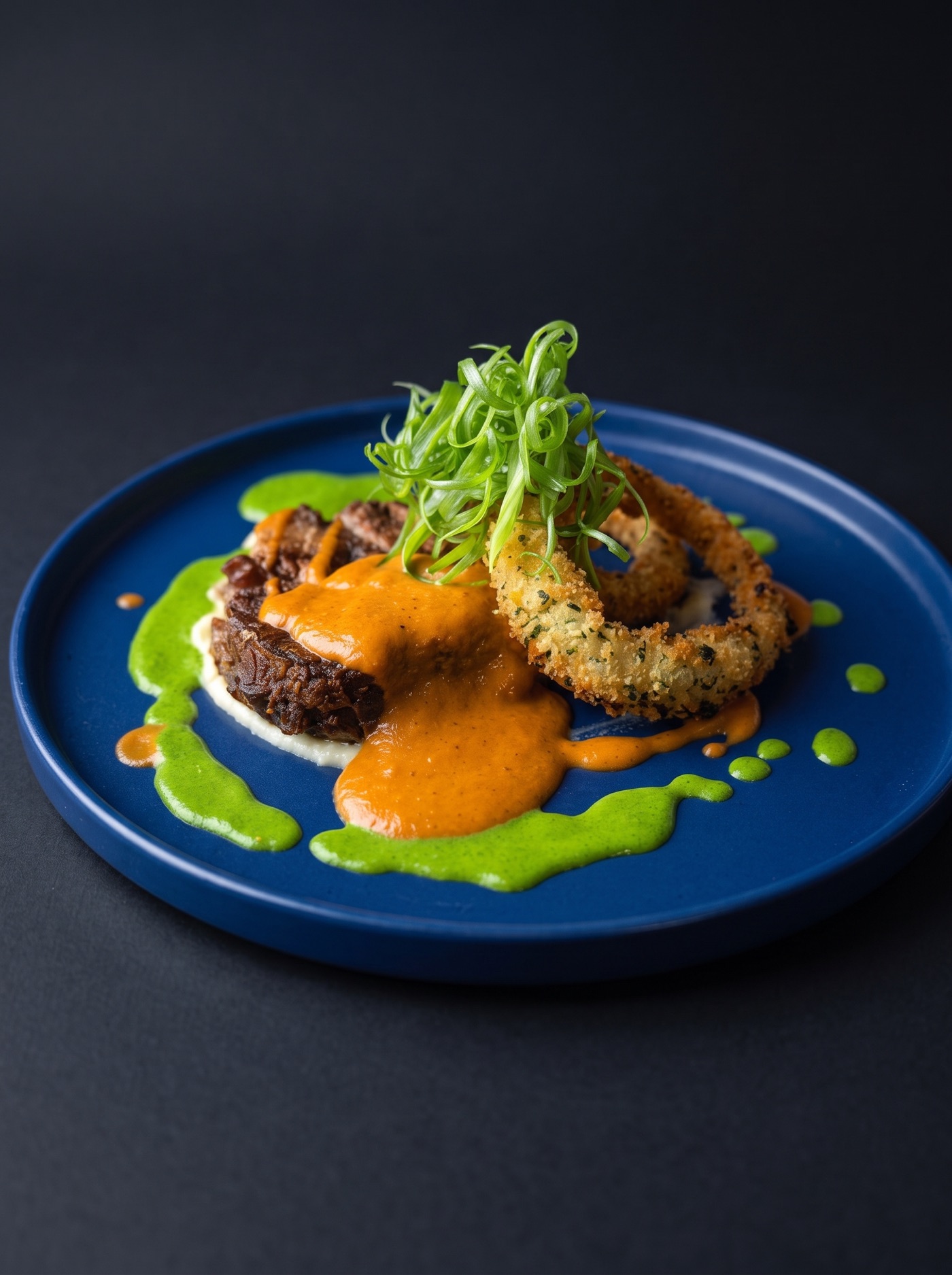 Braised short rib with romesco, herb purée and a panko-crusted onion ring, plated on cobalt